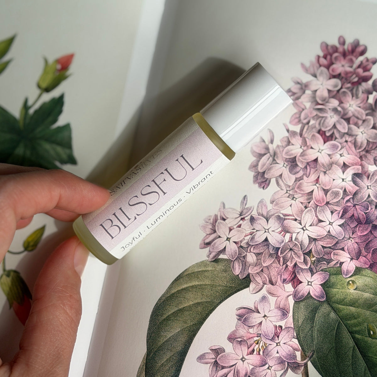 A hand holds a clear glass roll-on bottle labeled 'BLISSFUL' with a white cap, positioned over an open book featuring a botanical illustration of pink lilac flowers and green leaves.