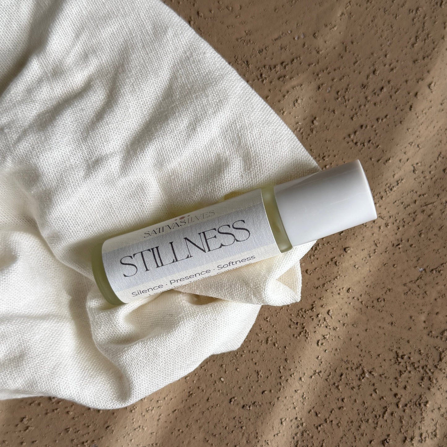 A small white cylindrical bottle labeled 'STILLNESS' by Saphirose, resting on a crumpled white fabric over a textured beige surface.