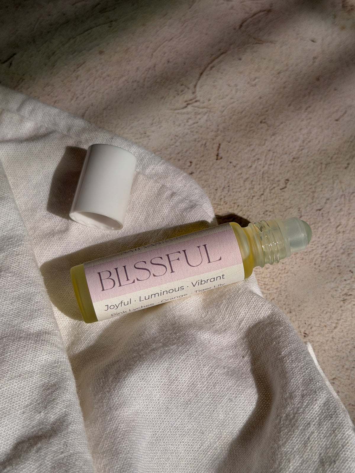 A roll-on bottle of Blissful essential oil with a pale pink label and white cap, resting on a textured beige linen fabric under warm, directional sunlight.