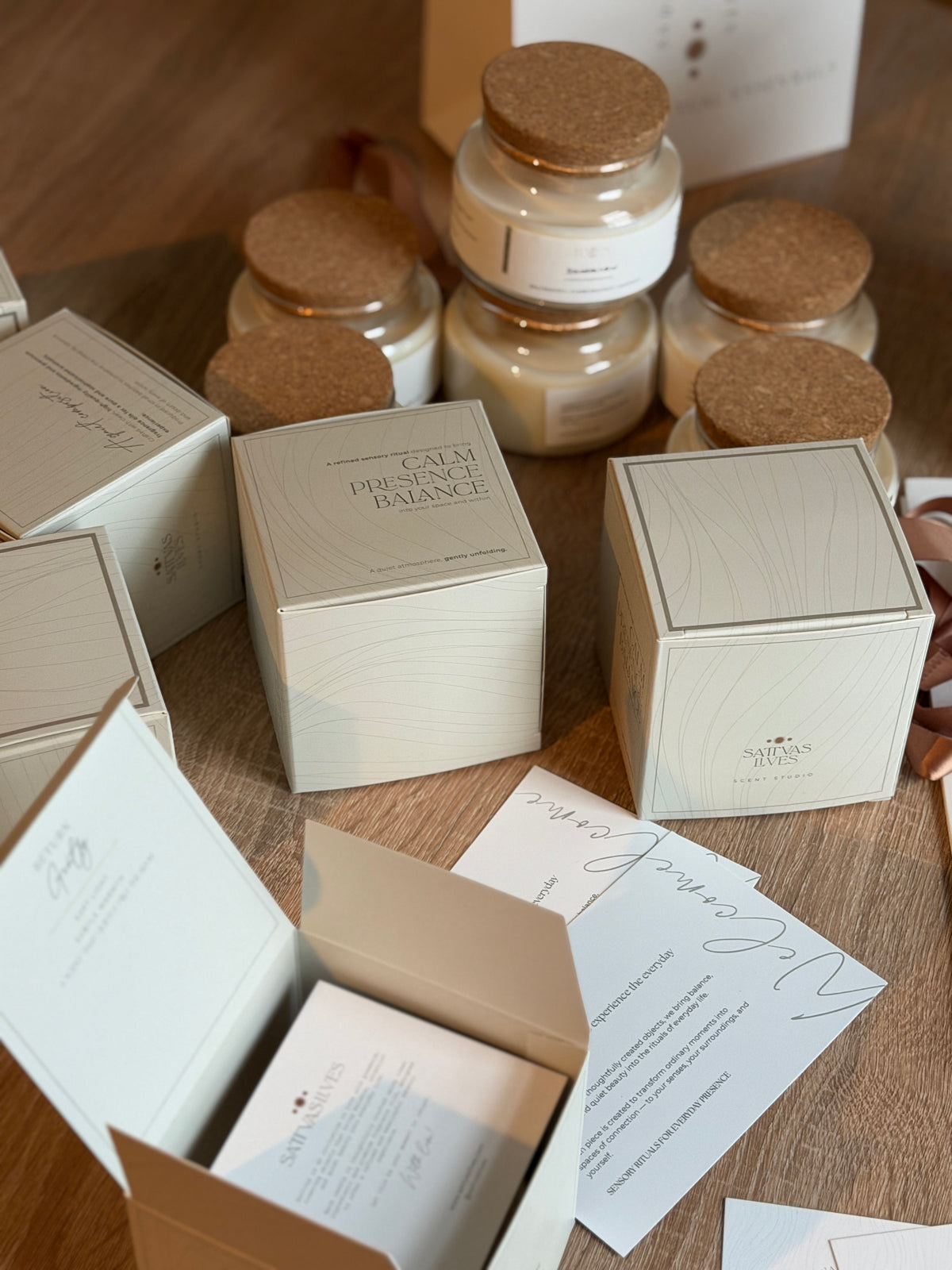 A styled flat-lay of a candle product line showing cream-colored packaging boxes, open boxes with printed cards, and several glass jars with cork lids containing creamy candles, all arranged on a wooden surface.