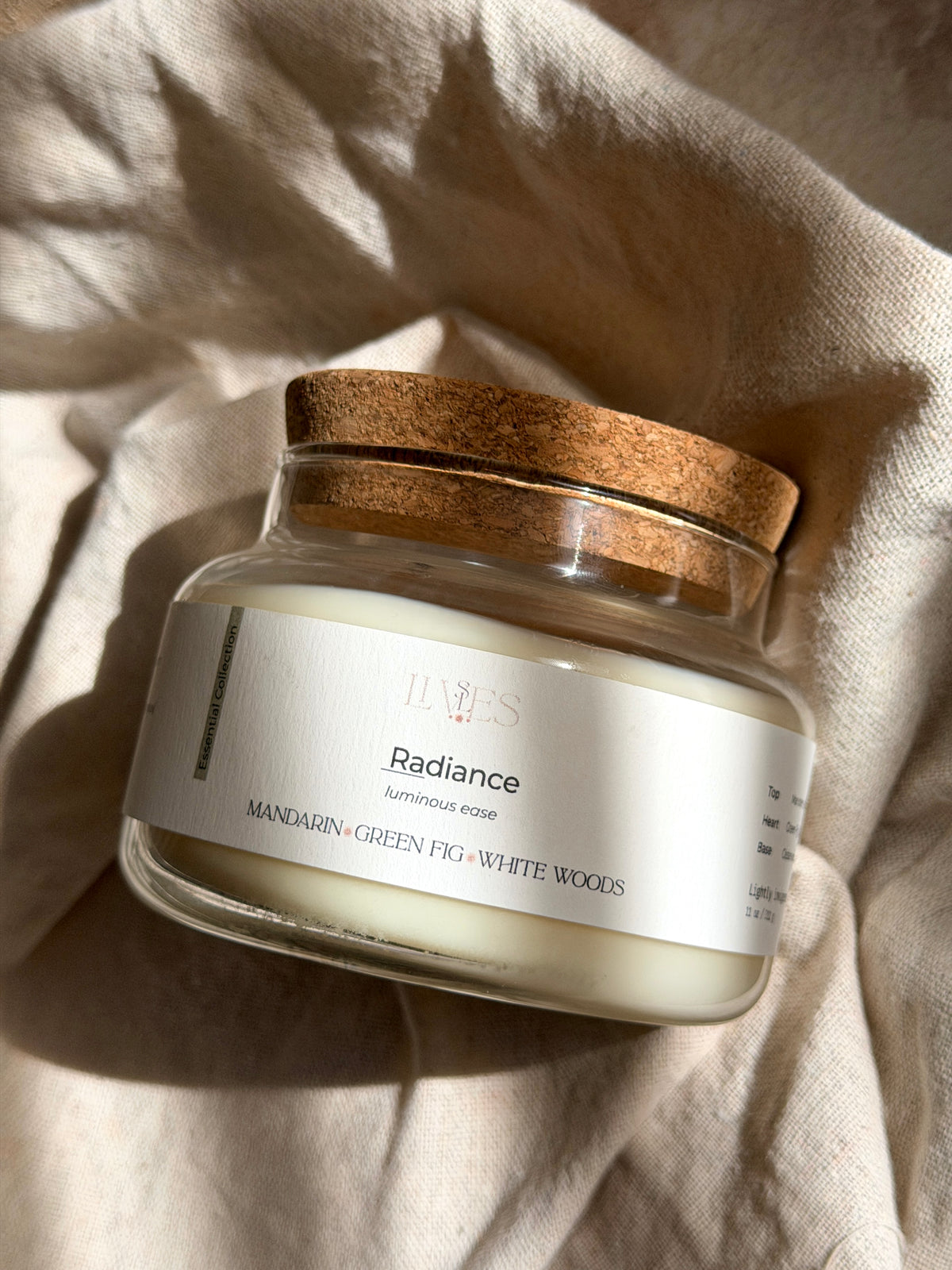 A glass jar candle with white wax and a natural wood lid, labeled 'Radiance' by JANE'S, resting on a textured beige linen fabric under warm sunlight.