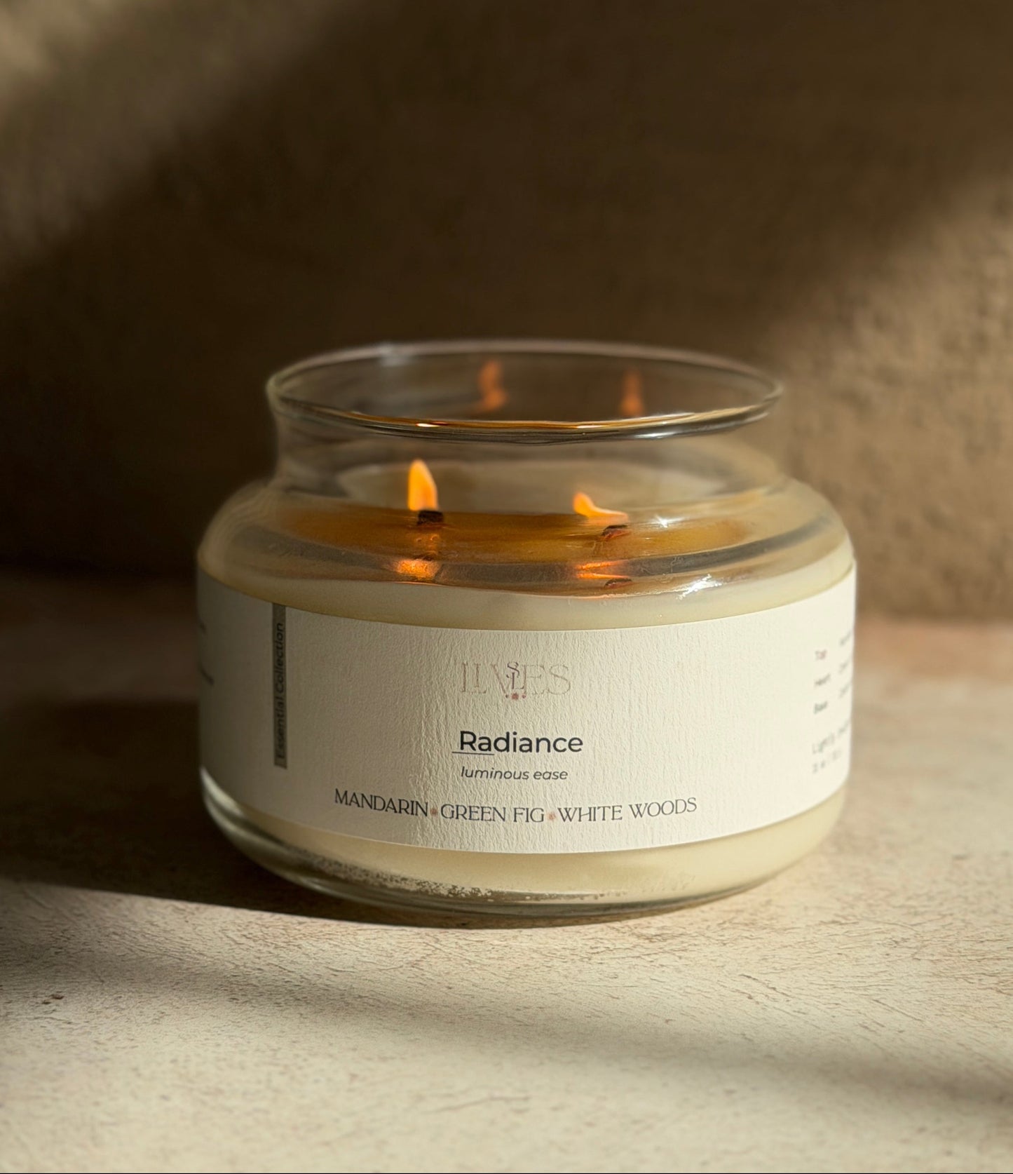 A lit candle in a clear glass jar with a cream-colored label, set on a textured beige surface with dramatic diagonal shadows.